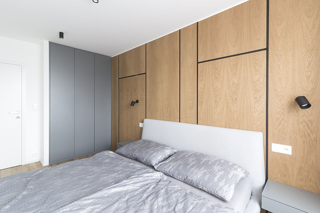 Design ložnice v kombinaci šedé barvy a masivního dubu, který tvoří obklad stěny, doplňují černá přísazná svítidla.