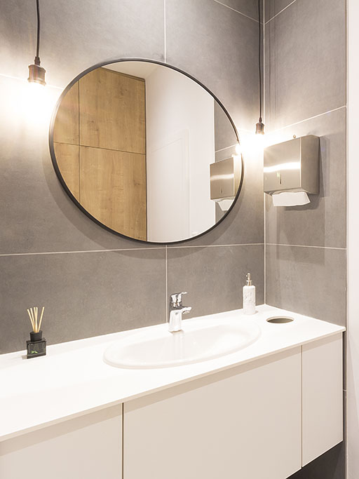 Design koupelny v interiéru kanceláří se zapuštěným umyvadlem a integrovaným košem na použité papírové útěrky doplněný o kulaté zrcadlo v černém rámu a závěsná svítidla.