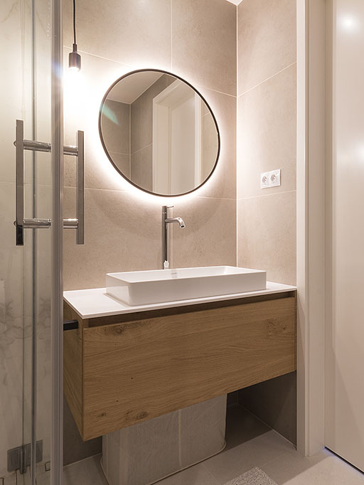 Interiérový design koupelny s dubovou skříňkou s umyvadlem a osvětleným kulatým zrcadlem.